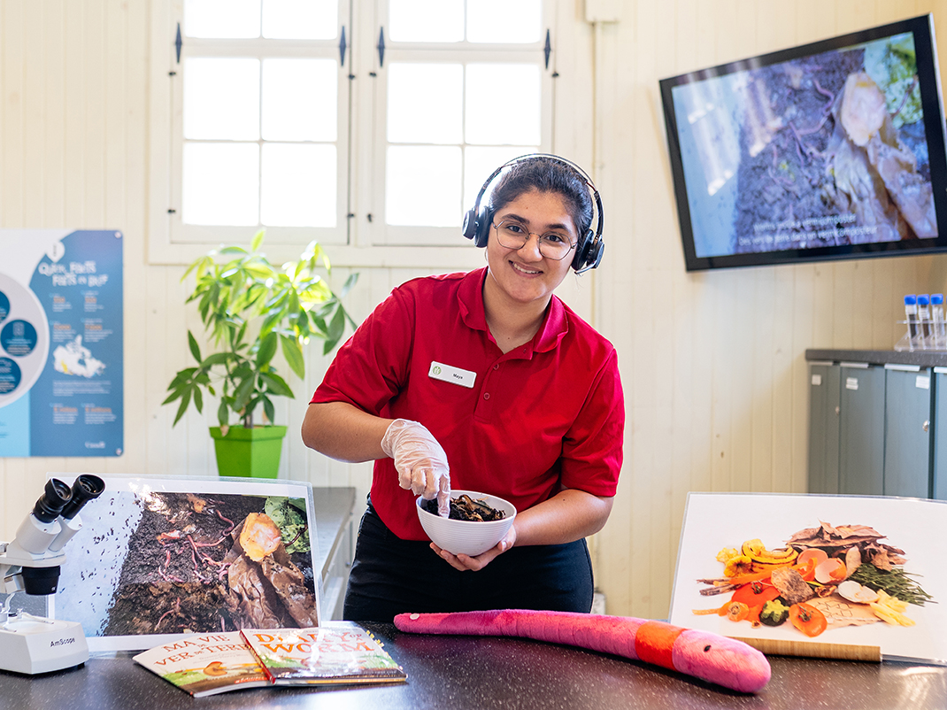 A museum employee with headphones demonstrates a bowl of soil, surrounded by plants, images and scientific diagrams.