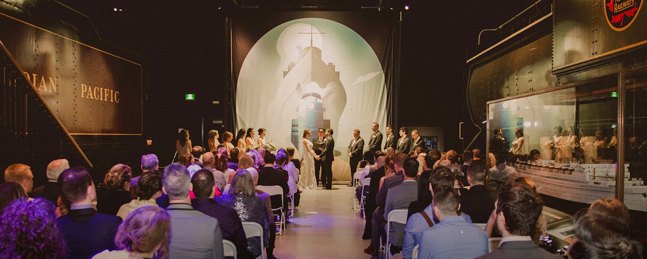 The scene of a wedding in between two trains at the Canada Science and Technology Museum. The bride and groom are holding hands at the front of the room while multiple attendees watch from white fold-out chairs in rows across the room. The bridesmaids and groomsmen are lined up at the front of the room. Above the officiant is a large white cloth featuring the faint outlines of a steamboat.