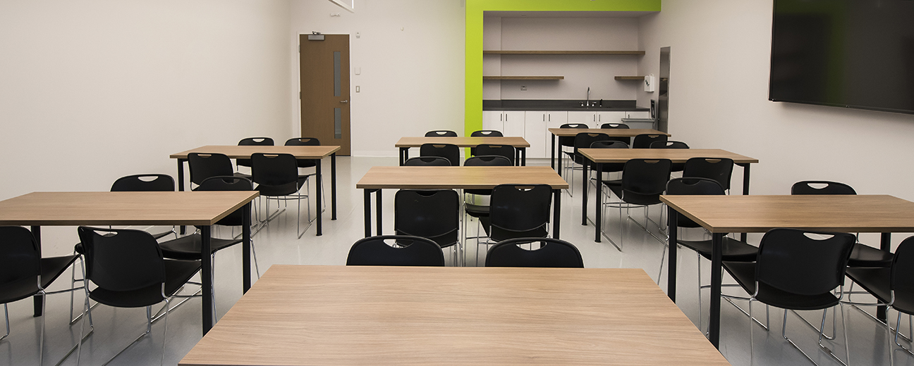 A spacious classroom with a TV on one side and eight desks with four chairs each arranged around the room. Another wall has a sink and a few shelves.