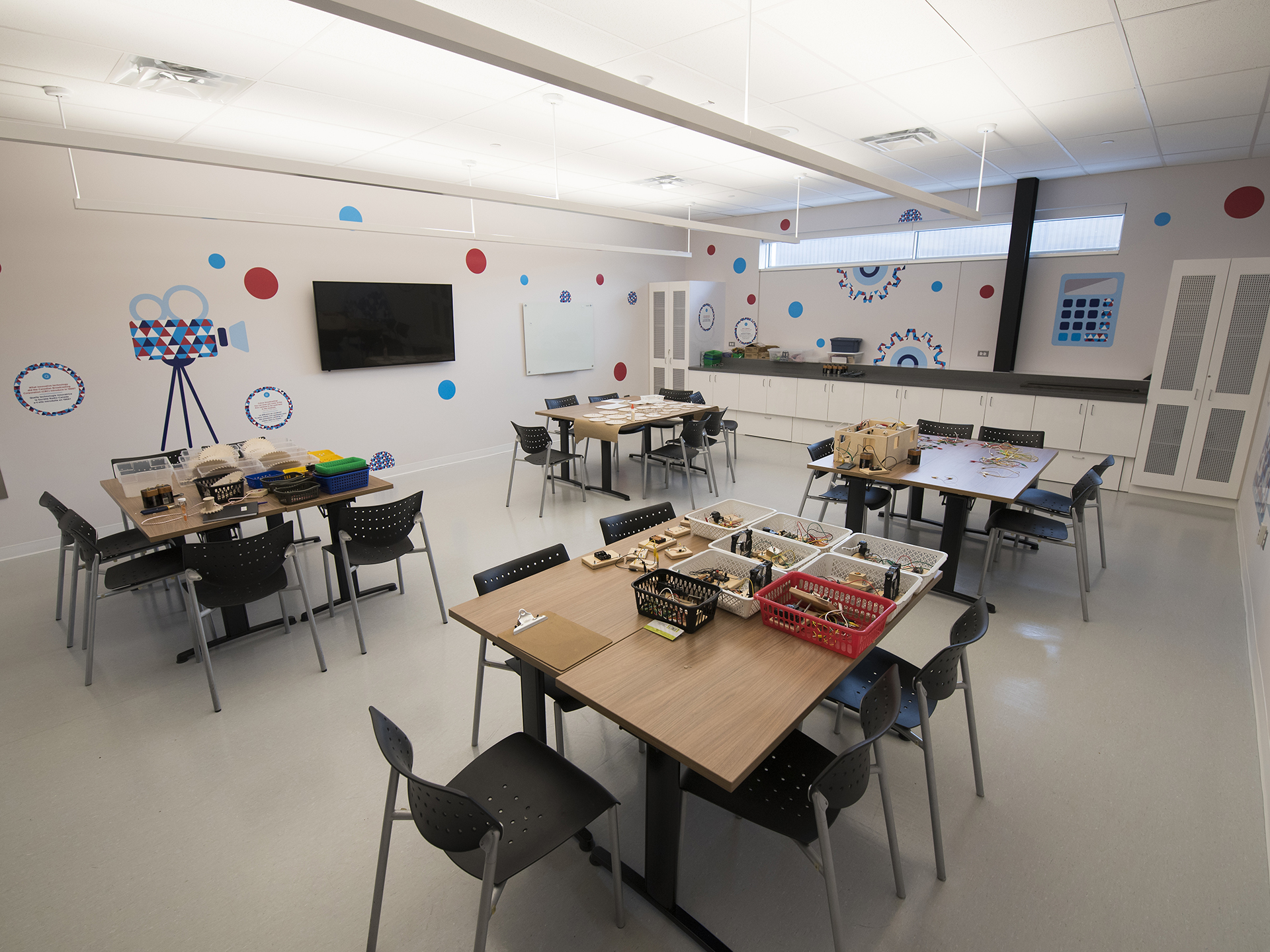 A spacious classroom with eight tables arranged in groups of two with five to six chairs at each table. A TV is displayed on one side of the room and the walls are decorated with colourful patterns including red and blue polka dots, gears, and the outline of a videocamera.