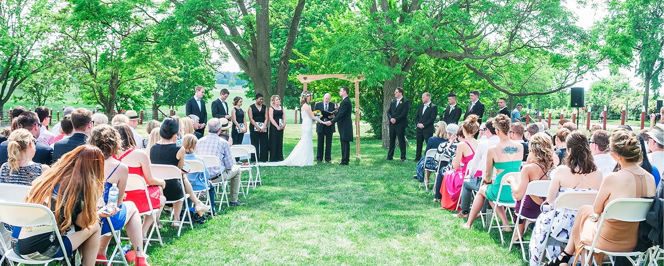 Beautiful outdoor wedding ceremony on a lush green lawn under large trees. Rows of people are sitting on white foldout chairs and a bride and groom are standing at the front under an arch with an officiant.