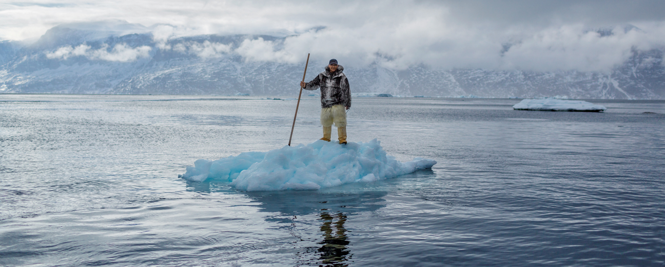 A man wearing a thick jacket and holding a spear standing on a small island of ice surrounded in all directions by water.