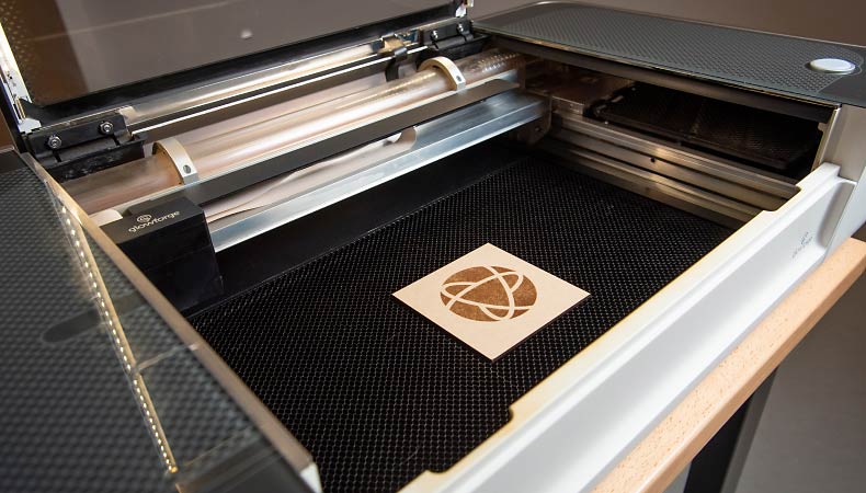 Close up of a laser cutter, and on the cutter there is a small wooden square with a circular logo on it.