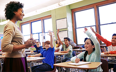 A classroom of smiling school-aged children are seated at their desks while raising their hands to answer a question posed by their teacher, who stands at the front of the class.