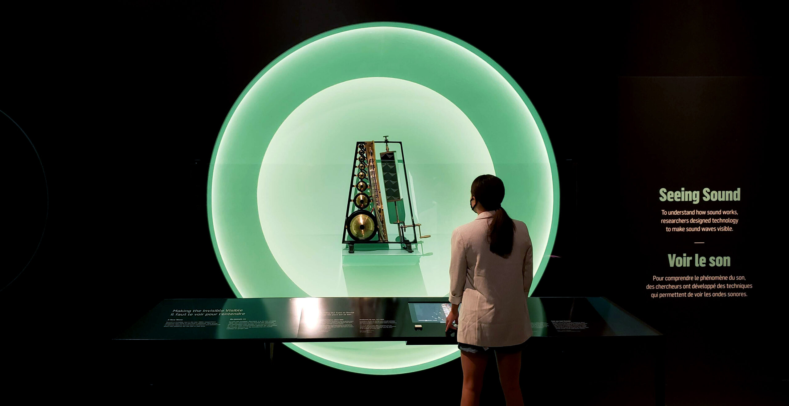 La silhouette d’une femme se découpe devant une vitrine circulaire illuminée où l’analyseur de son Koenig est exposé. Le mur porte le titre « Voir le son ».