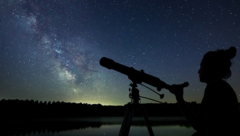 The silhouette of a woman standing by a telescope under a dark blue starry sky, with a serene lake and tree-lined shore in the background.