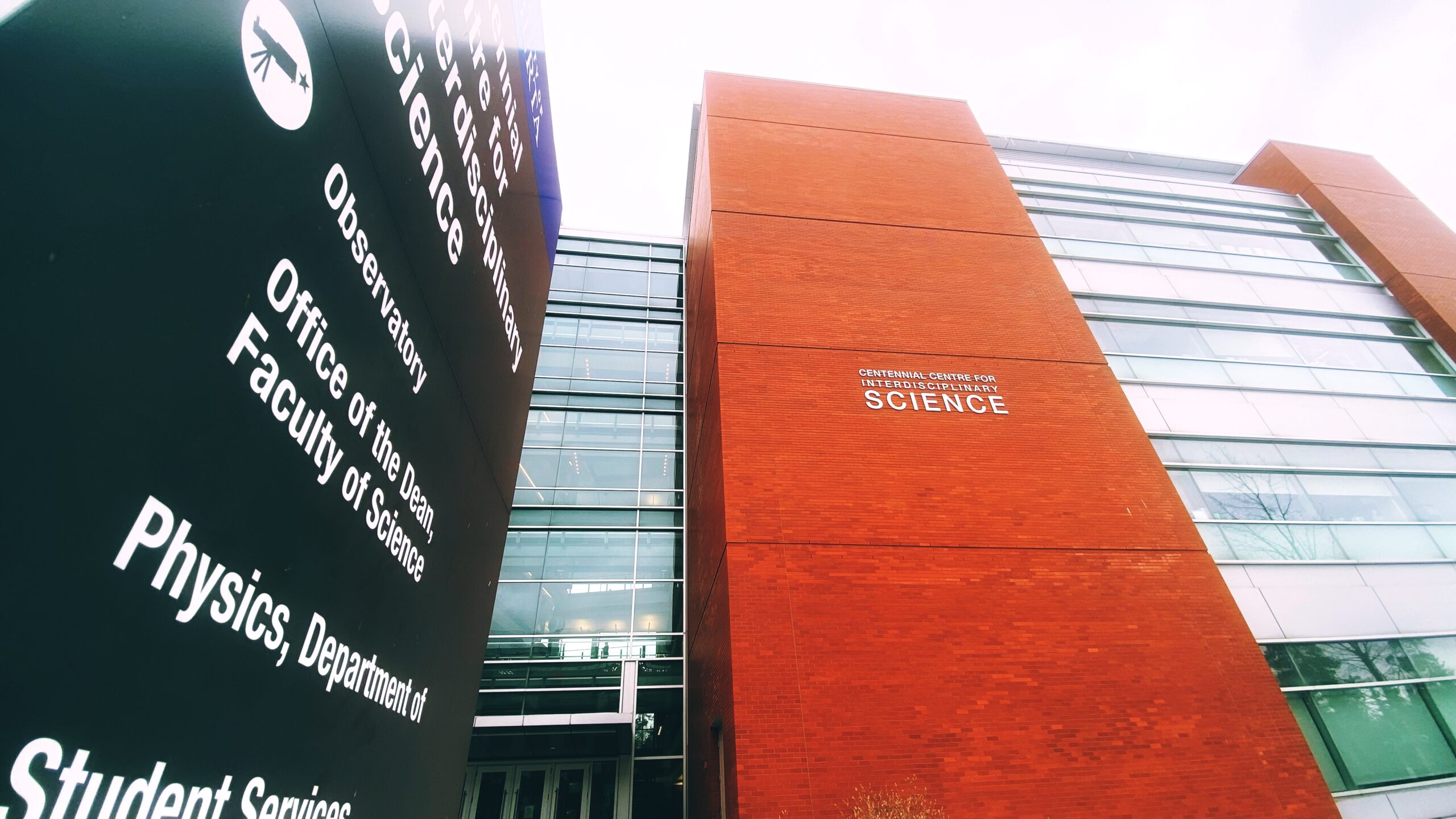 Vue en contre-plongée d’un immeuble de l’Université de l’Alberta. À gauche, un panneau affiche plusieurs indications, comme « Physics, Department of » (Département de physique) en lettres blanches. À droite, les mots « Centennial Centre for Interdisciplinary Science » (Centre Centennial de sciences interdisciplinaires) sont directement apposés sur la façade de l’immeuble, sur un mur en briques rouges de plusieurs étages bordé de fenêtres de chaque côté.