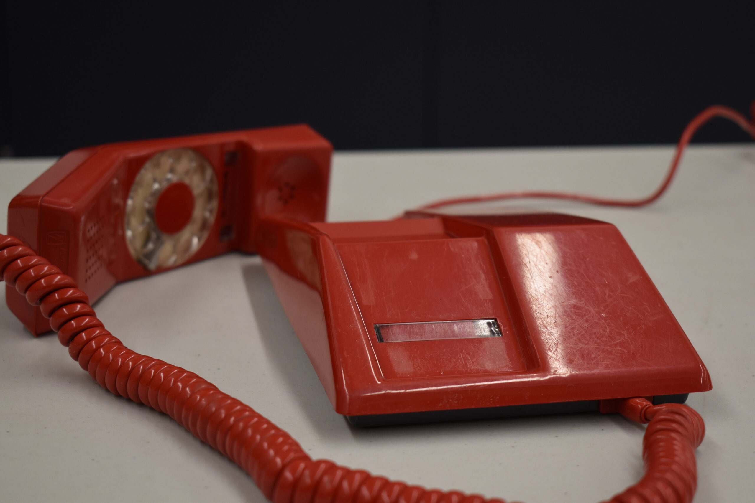 Un téléphone en plastique rouge posé sur une table gris clair, avec fond noir. Le combiné du téléphone, dans lequel se trouve le cadran rotatif, est posé sur la table à côté de la base de l’appareil, et est relié à celle-ci par un cordon rouge en spirale.