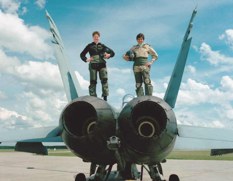 Two servicewomen in flight suits pose confidently atop a fighter jet at an airstrip on a clear blue day.