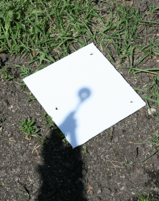 Une photographie montre l'ombre d'un clinomètre se reflétant sur une feuille de papier blanc posée sur le sol. Un petit trou de lumière solaire apparaît à travers le tube du clinomètre.