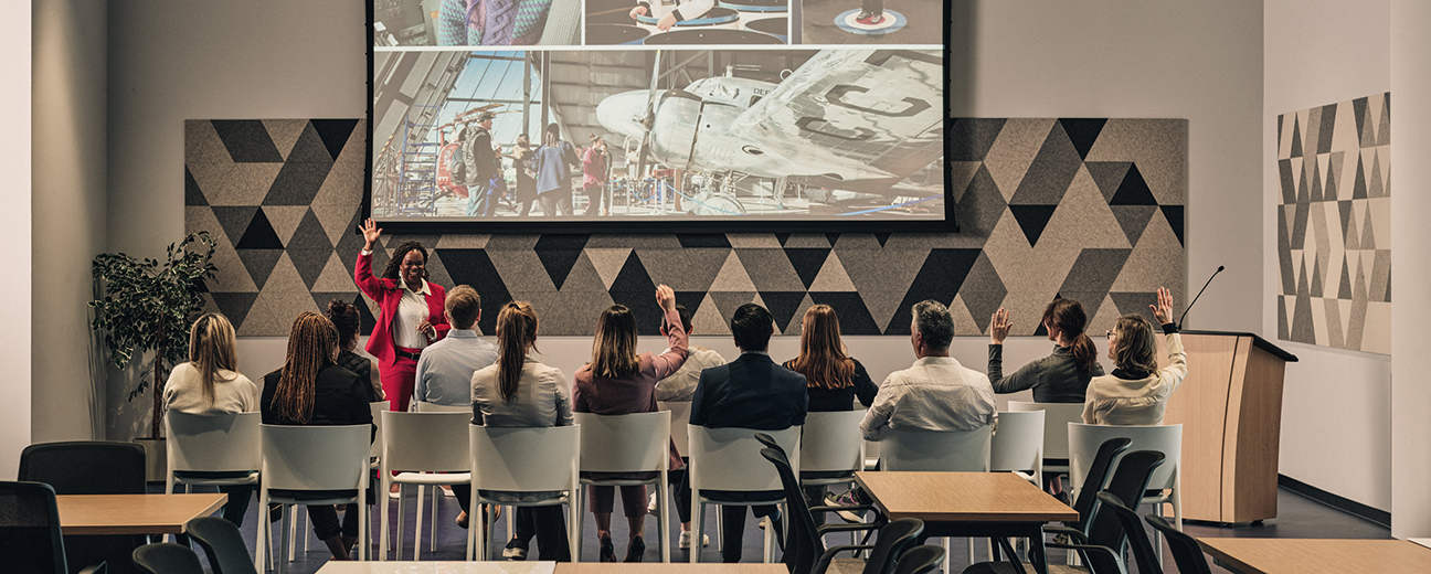 A presenter in a bright red suit engages an audience seated in a modern conference room, with a large screen displaying aviation-themed images in the background. Some attendees have their hands raised, participating actively in the session.