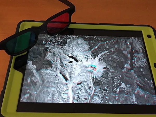 A pair of 3D glasses, with one red lens and one blue lens, leaning on an iPad in a green case. The iPad has an anaglyph satellite image of Mount St Helens, for viewing with the glasses, on its screen.