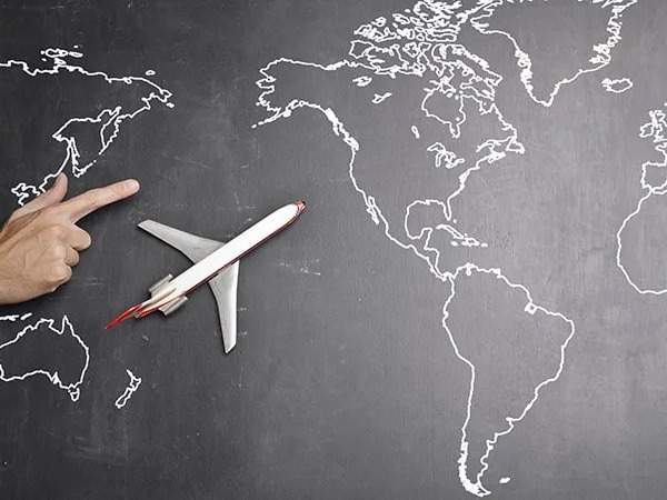 A model commercial aircraft against a blackboard with a map of the world drawn in white chalk. Beside the aircraft is a hand with a finger pointing, indicating the direction the aircraft is flying.