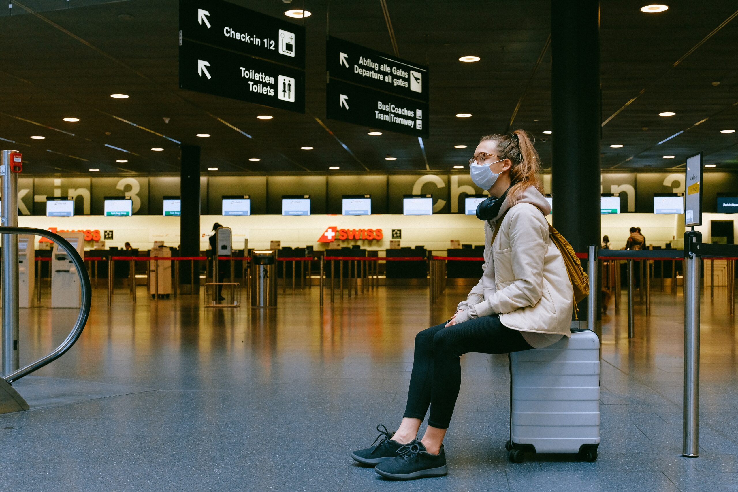 Dans une aérogare vide, une jeune femme est assise sur sa valise et porte un masque médical; elle a des écouteurs autour du cou.