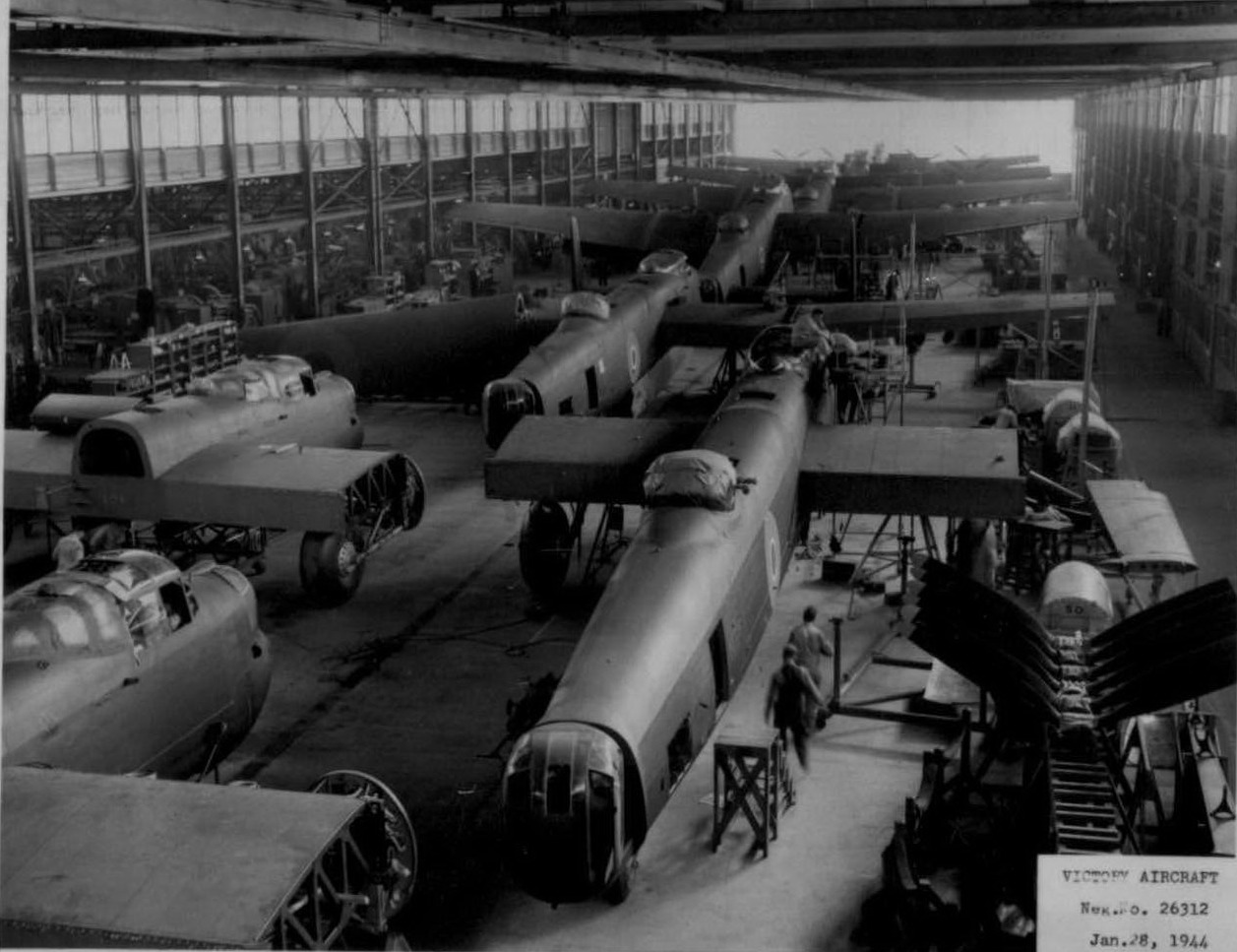 A black and white photograph of approximately 10 airplanes under construction in a large factory. Two workers are visible in the foreground.