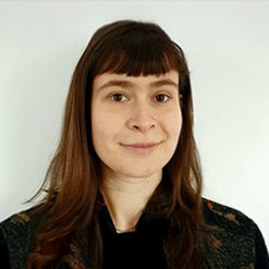 Person with brown eyes, long brown hair and bangs smiling in front of a white wall. The person is wearing a dark floral shirt with orange and pink flowers.