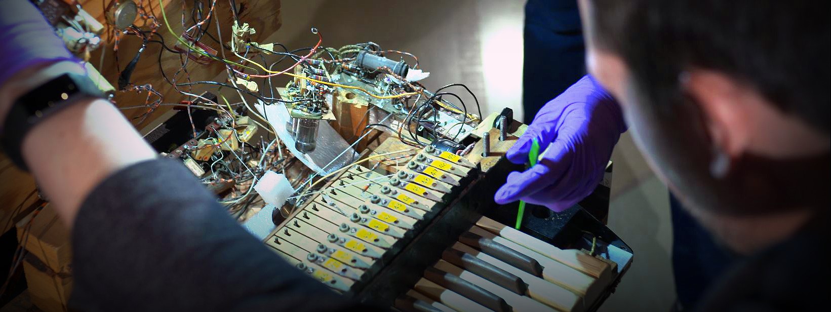 A person looking down at the inside of the electronic sackbut. A purple gloved hand is reaching towards a tangle of wires wires.