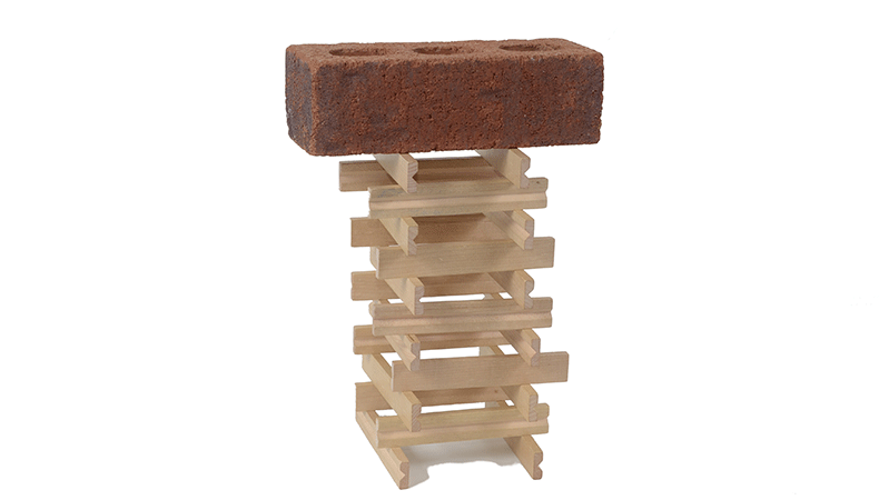 Wooden sticks are stacked, two on each layer and alternating direction in each layer to form a square based tower with a hollow centre. A brick sits atop the tower.