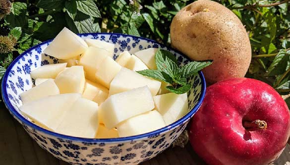 Diced pieces of apple and potato in a bowl. Sprigs of mint, an apple and a potato sit beside the bowl.