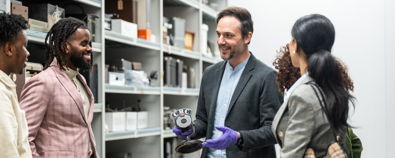 A museum professional wearing a dark grey blazer and purple gloves holds an old CBC microphone. There are four visitors listening attentively and the setting is a museum storage area, with white shelves and various artifacts visible in the background.