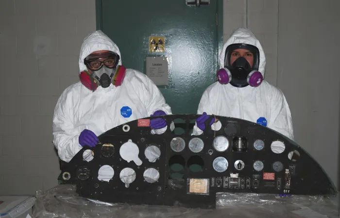 Deux conservateurs du musée en tenue complète de protection individuelle posent côte à côte et tiennent à la verticale un panneau de commande d’avion de couleur noire. L’artefact est posé sur une feuille de plastique qui recouvre une table.